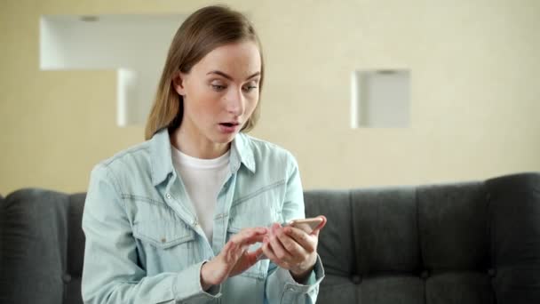 Mujer feliz sosteniendo el teléfono inteligente y haciendo un trato exitoso mientras trabaja desde casa. Mujer ganando mucho dinero y regocijándose. Dinero cayendo desde arriba. — Vídeos de Stock