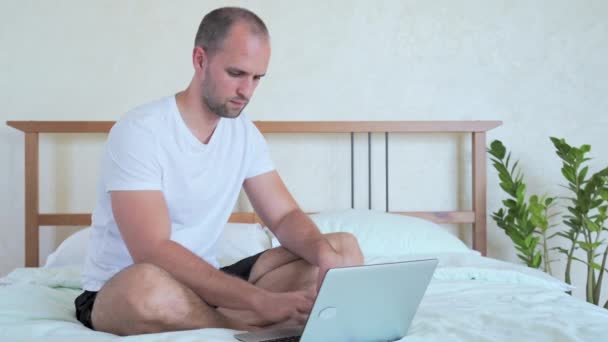 Homem está trabalhando com um laptop, sentado na cama . — Vídeo de Stock