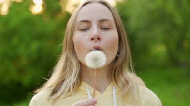 Een mooie jonge vrouw staat in het bos en blaast een paardebloem. Op straat. Geniet van de natuur. Een gezond, lachend meisje op een zomergazon. Allergievrij concept — Stockvideo