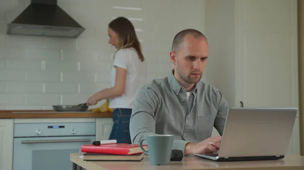 Mujer cocinando en la cocina y el hombre trabajando con el ordenador portátil en la mesa en casa — Foto de Stock