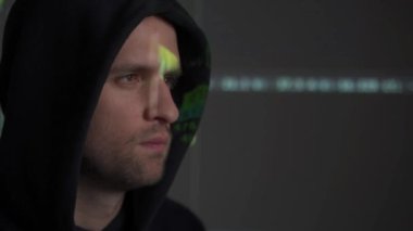 Yeşil kod karakterleri karanlık bir ofiste yüzünü yansıtırken, erkek hacker bilgisayar üzerinde çalışıyor..