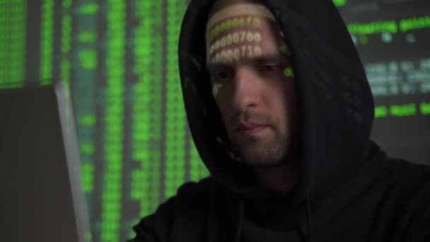 Hacker masculino trabalhando em um computador, enquanto personagens de código verde refletem em seu rosto em uma sala de escritório escuro . — Vídeo de Stock