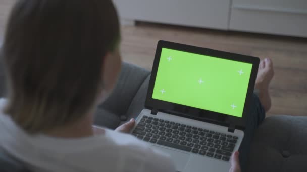 Mujer en casa sentada en un sofá tapones en una computadora portátil con pantalla verde simulada. chica usando la computadora, navegar a través de Internet, viendo contenido — Vídeo de stock