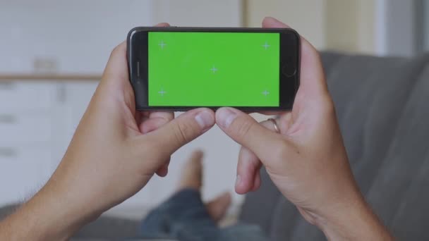 Hombre en casa acostado en un sofá usando con Smartphone de pantalla verde en modo paisaje horizontal. Chica usando viendo videos . — Vídeos de Stock