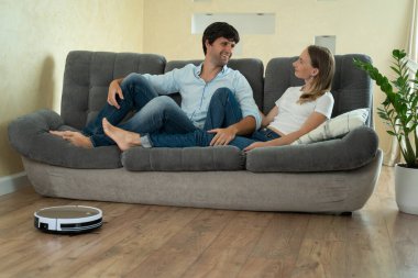 Genç çift koltukta dinlenirken otomatik elektrikli süpürge oturma odasında ev işlerini yapıyor. Ev robotu kavramı
