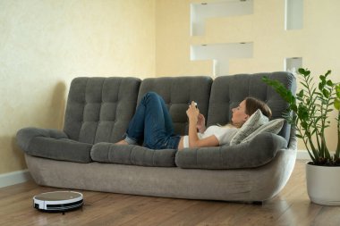 Akıllı telefon kullanan çekici bir kız. Robotik elektrikli süpürge ile temizlik yapıyor.