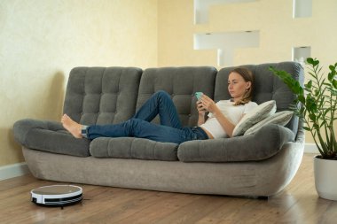 Akıllı telefon kullanan çekici bir kız. Robotik elektrikli süpürge ile temizlik yapıyor.