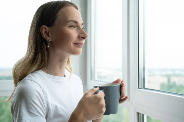 Mujer mirando por la ventana disfrutando de un nuevo día fresco sintiéndose descansado y tomando café en casa. Moción lenta — Foto de Stock