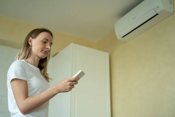 Vrouw in haar moderne appartement in de woonkamer. Ze zet de airco van de afstandsbediening aan. Klimaatbeheersing thuis met splitsysteem. — Stockfoto