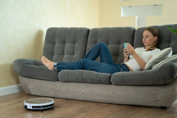 Chica atractiva utilizando el teléfono inteligente mientras que la aspiradora robótica casa de limpieza — Foto de Stock