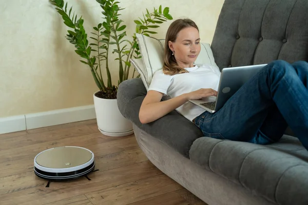 Az okos porszívó kitakarítja a házat. Egy nő számítógéppel a kanapén fekszik, és egy robot porszívó takarít.. — Stock Fotó