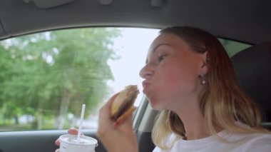 Hamburger yiyen aç bir kadın arabada oturuyor, abur cubur yiyor.