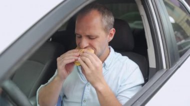 Adam arabasını sürerken hamburger yiyor.