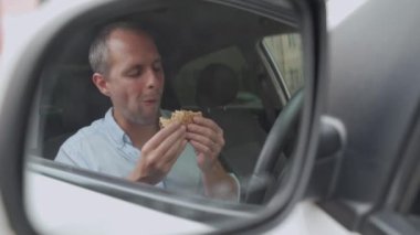 Adam arabasını sürerken hamburger yiyor.