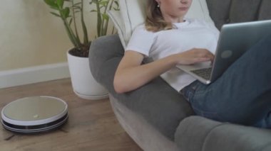 Akıllı elektrik süpürgesi evi temizliyor. Bilgisayarlı bir kadın kanepede uzanıyor ve elektrikli süpürge temizliyor..