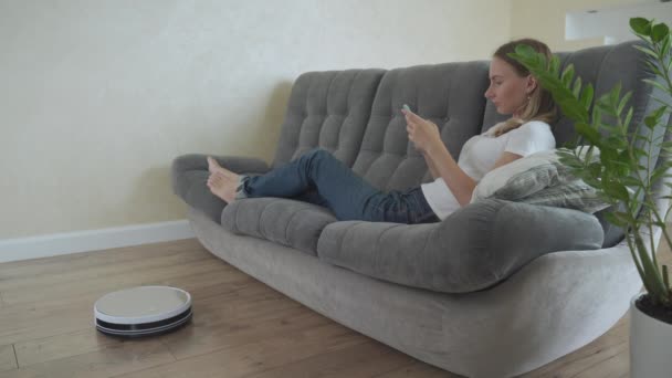 Fille attrayante en utilisant smartphone tandis que robot aspirateur nettoyage maison — Video