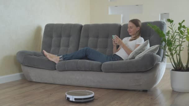 Привлекательная девушка с помощью смартфона в то время как робот пылесос уборка дом — стоковое видео