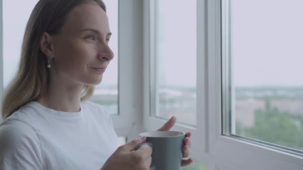 Жінка, дивлячись у вікно, насолоджується новим днем, відчуваючи відпочинок і п'є каву вдома. Повільний рух — стокове відео