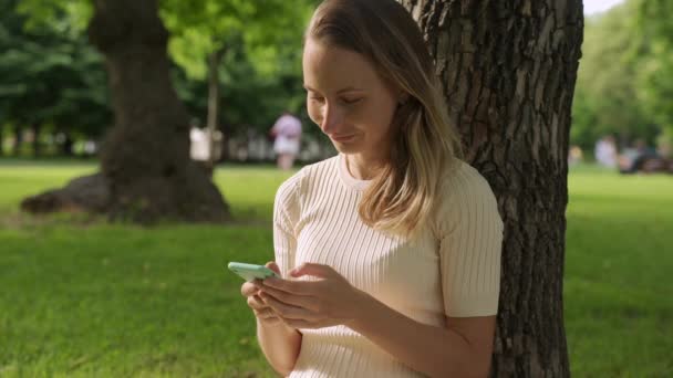 Dekat dengan seorang wanita muda yang cantik, yang menggunakan aplikasi di ponsel pintarnya di taman. Berkomunikasi tentang ponsel, teknologi, aplikasi — Stok Video