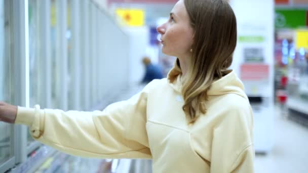 Женщина покупает замороженные продукты в супермаркете. Привлекательная молодая женщина стоит рядом с морозильной камерой и выбирает рыбу. Покупка продуктов питания в супермаркете. — стоковое видео