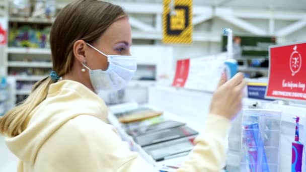 Женщина в маске выбирает электрическую зубную щетку в магазине — стоковое видео