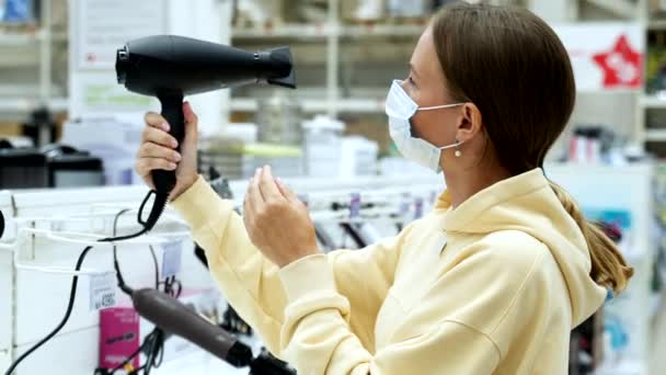 Una mujer enmascarada sosteniendo un secador de pelo. En el fondo, estantes con electrodomésticos. El concepto de comprar electrodomésticos para la belleza — Vídeos de Stock