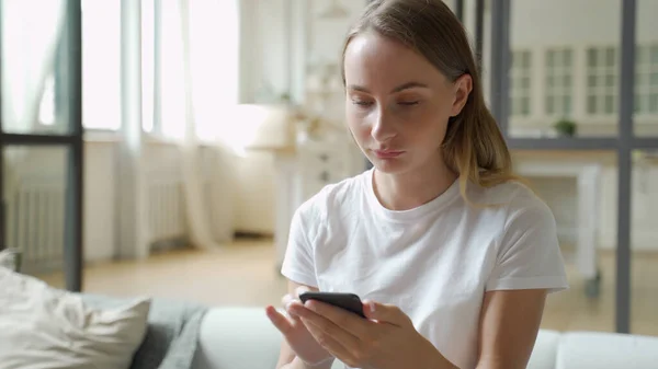 Jovem mulher sorrindo usando telefone celular enquanto se senta no sofá em casa — Fotografia de Stock