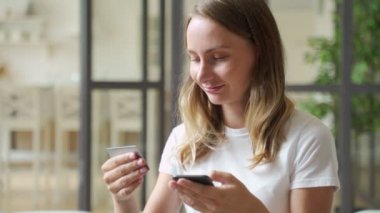 Kredi kartı ve akıllı telefon tutan bir kadın. Kanepede oturan genç bir kadın evde para harcıyor, sipariş veriyor, internet, ticari hizmetler ve çevrimiçi ödeme kavramını kullanarak satın alıyor.