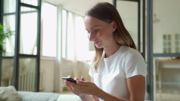 Jovem mulher sorrindo usando telefone celular enquanto se senta no sofá em casa — Vídeo de Stock