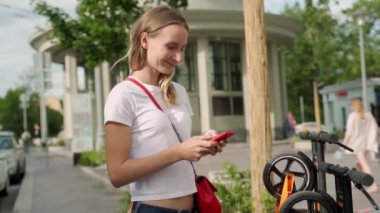 Modern şehir caddesinde park yerinde dikilirken akıllı telefon kullanan bir kadın elektrikli scooter kiralıyor.
