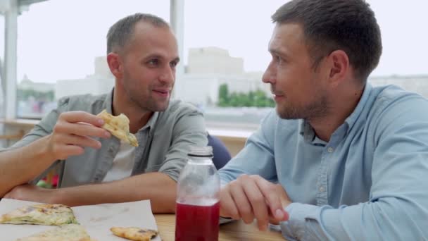 Два друга едят пиццу и разговаривают в кафе — стоковое видео