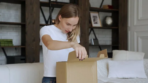 Woman sit on couch in living room open cardboard box shopping online at home, happy female unpack parcel order purchase goods on Internet.