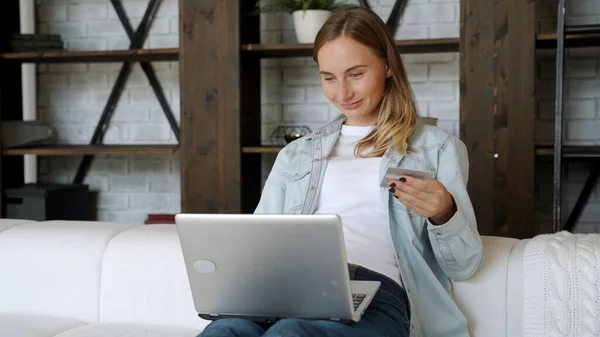 Mujer con tarjeta de crédito y el uso de la computadora portátil, sentado en el sofá en casa, las compras de clientes jóvenes en línea en la tienda de Internet — Foto de Stock