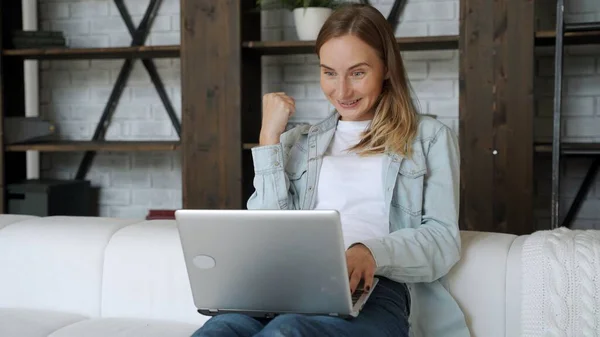 Όμορφη νεαρή γυναίκα χρησιμοποιεί ένα φορητό υπολογιστή και χαμογελώντας, ενώ κάθεται στον καναπέ στο σπίτι — Φωτογραφία Αρχείου