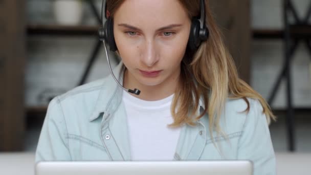 Mujer joven con un auricular que trabaja en línea en la computadora mientras está sentada en casa. Concepto de estudiar o trabajar desde casa en línea — Vídeos de Stock