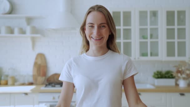 Joyeux jeune femme avec des bretelles sourit dans le fond de sa cuisine, en regardant la caméra — Video