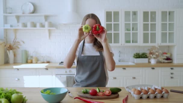 Krásná žena si hraje se dvěma papričkami v kuchyni. Emocionální žena dělá obličeje se zelenou a červenou paprikou — Stock video