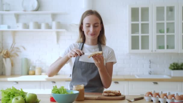 Счастливая улыбающаяся женщина на кухне готовит сэндвич с арахисовым маслом — стоковое видео