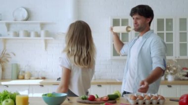 Genç çift ellerinde mutfak eşyalarıyla gülüyor birlikte şarkı söylüyor müzik eşliğinde dans ediyor mutfakta yemek pişiriyor..