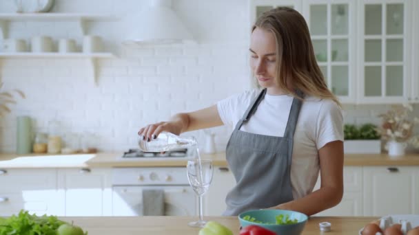 Gros plan d'une femme versant de l'eau fraîche dans un verre à partir d'une bouteille dans sa cuisine. — Video