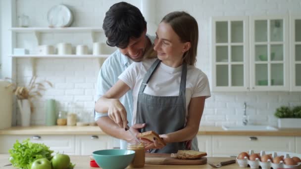 Κοντινό ευτυχισμένο ζευγάρι που μαγειρεύουν μαζί στο σπίτι. Όμορφος άντρας και γυναίκα διασκεδάζουν. Ένα νεαρό ζευγάρι ετοιμάζει ένα σάντουιτς με φυστικοβούτυρο. — Αρχείο Βίντεο