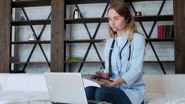 La mujer usa una bata blanca y un auricular mientras habla en una computadora portátil usando una aplicación de consulta de videollamadas en línea. Atención médica remota para un paciente remoto — Foto de Stock