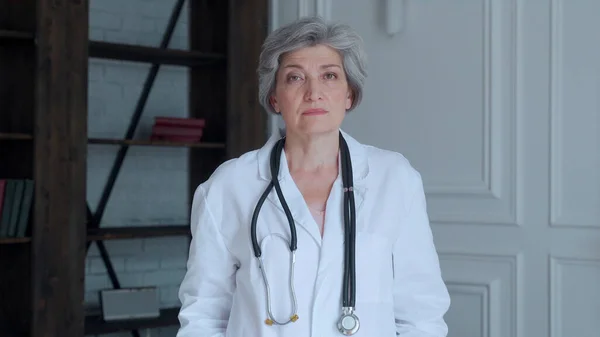 Portrait of an elderly doctor in a dressing gown, with a stethoscope on a light blue background of bookshelves — Stock Photo, Image