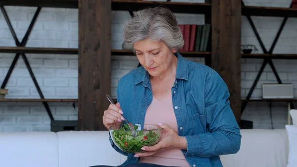 Пожилая женщина ест овощной салат, сидя дома на диване. — стоковое фото
