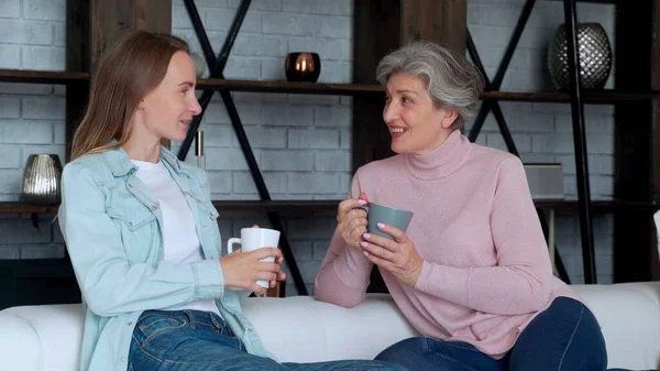 Madura madre y su hija adulta están tomando café, hablando y sonriendo mientras están sentados en el sofá en casa — Foto de Stock