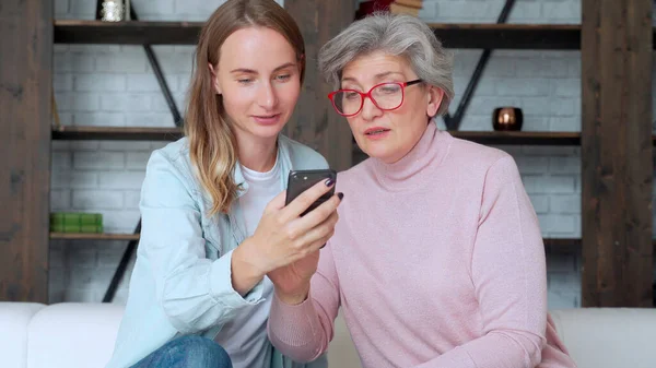 Elderly mother and an adult daughter are sitting on the couch at home, having fun using a smartphone together, watching videos on a mobile phone.
