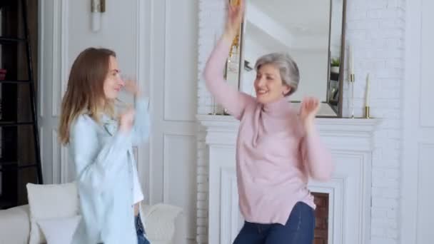 Ancianos madura madre de pelo gris se divierte con una hija feliz en casa. Alegre nieta baila a su música favorita con una feliz madre anciana — Vídeos de Stock