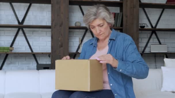 Äldre kvinna sitter på soffan i vardagsrummet öppen kartong handla online hemma. Leveranskoncept — Stockvideo