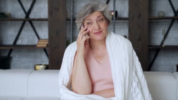 Una anciana está descansando en un sofá acogedor, hablando en un teléfono inteligente con amigos. Una anciana está charlando en un teléfono móvil con niños adultos. — Vídeo de stock