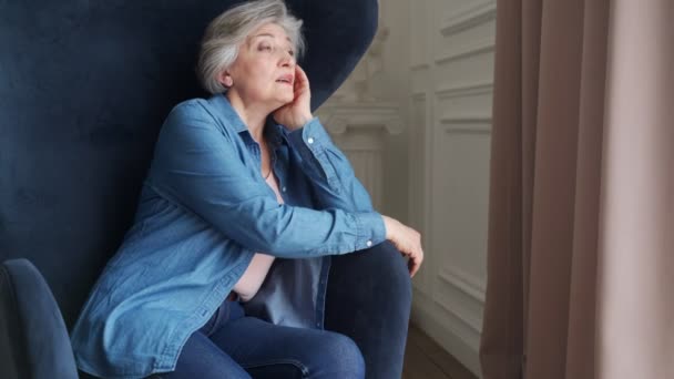 Äldre kvinna sitter ensam i en stol hemma och tittar ut genom fönstret. En pensionär vilar i vardagsrummet. — Stockvideo
